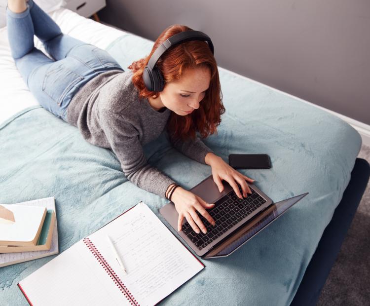 Regardant vers le bas sur l'étudiant féminin d'université utilisant des écouteurs se trouvant sur le lit fonctionnant sur l'ordinateur portatif